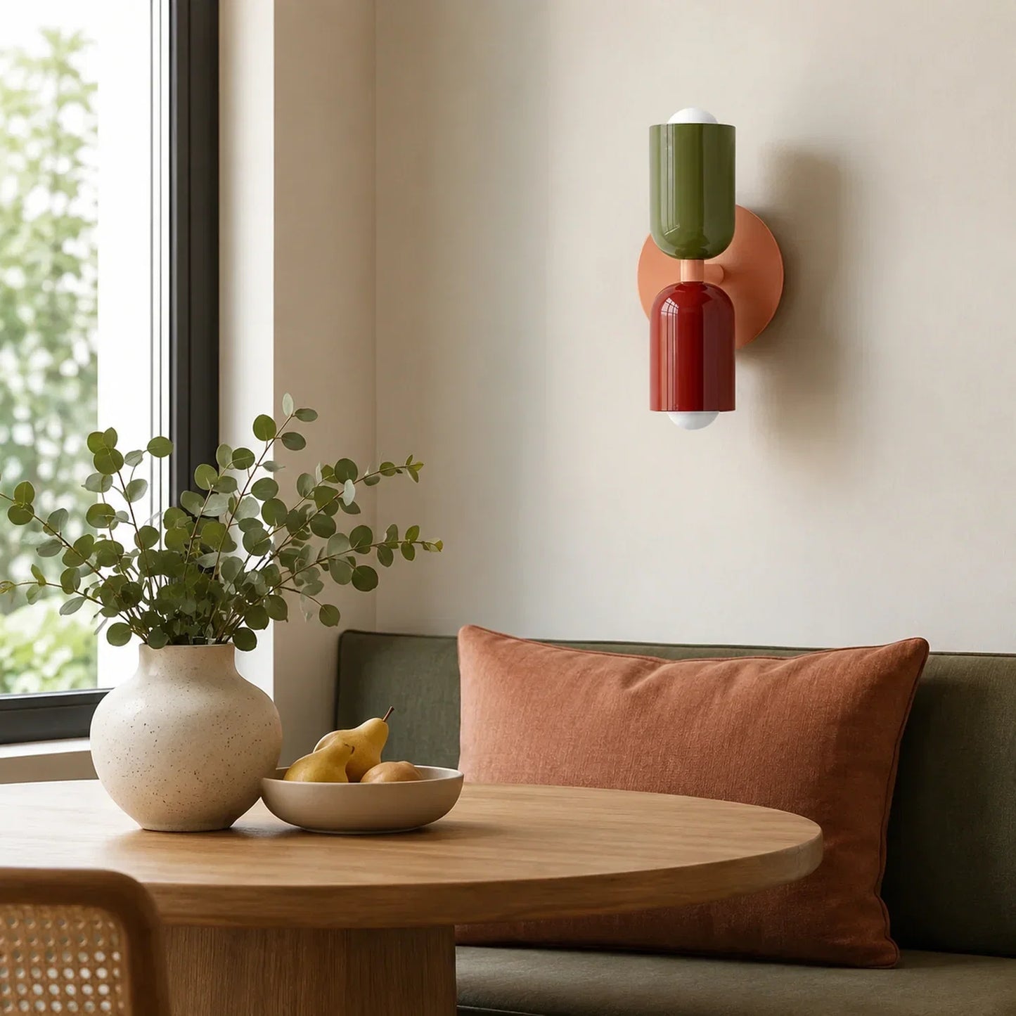 Contemporary dual-cylinder metal wall lamp with green and red shades mounted on a wall above a dining table with a vase, foliage and a bowl of pears.