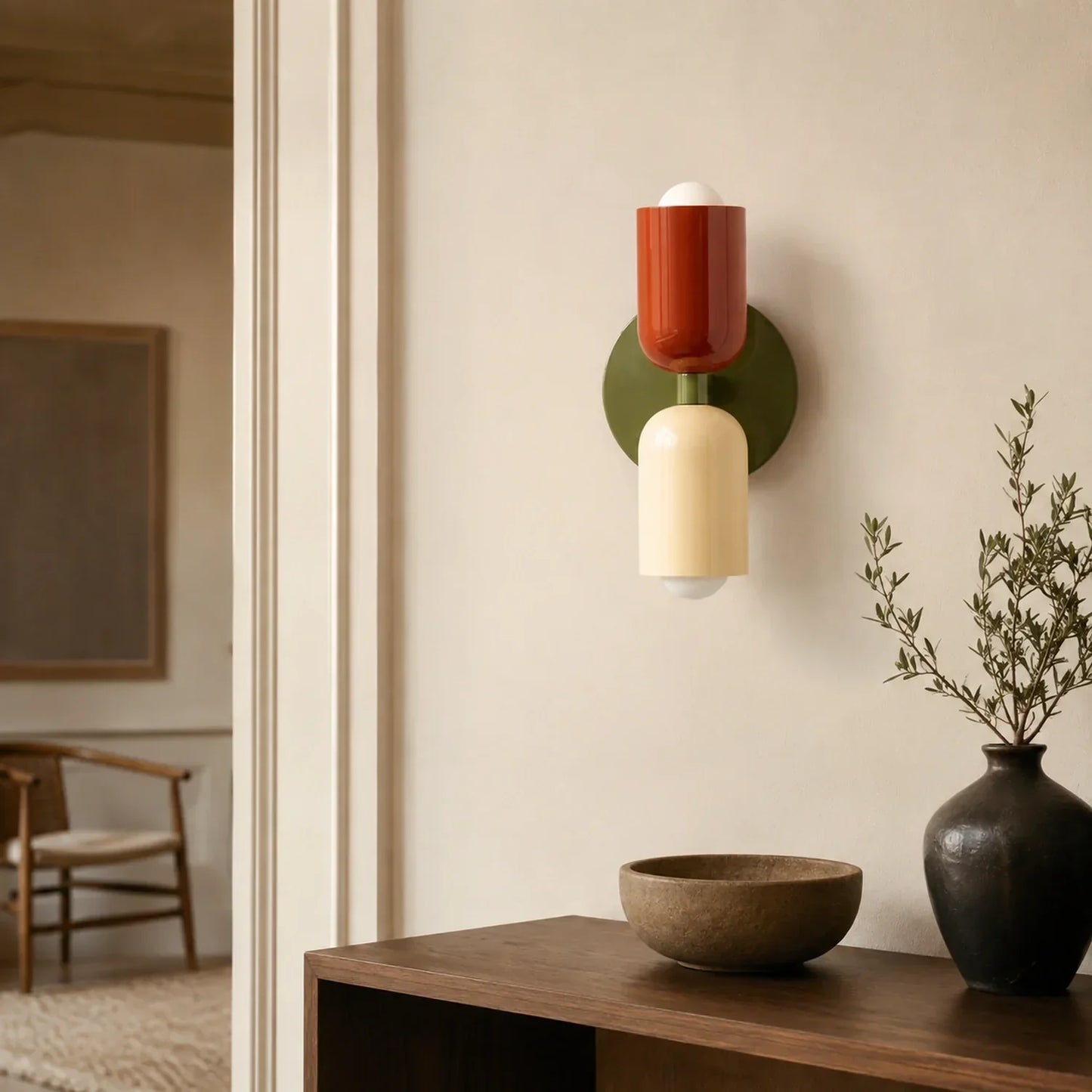 Contemporary dual-cylinder metal wall lamp featuring green, red, and cream accents, mounted on a neutral wall above a wooden console with a vase.
