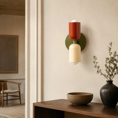 Contemporary dual-cylinder metal wall lamp featuring green, red, and cream accents, mounted on a neutral wall above a wooden console with a vase.