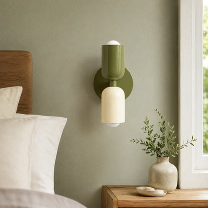 Contemporary dual-cylinder metal wall lamp in green and cream mounted on a sage green bedroom wall above a wooden bedside table with a small plant.