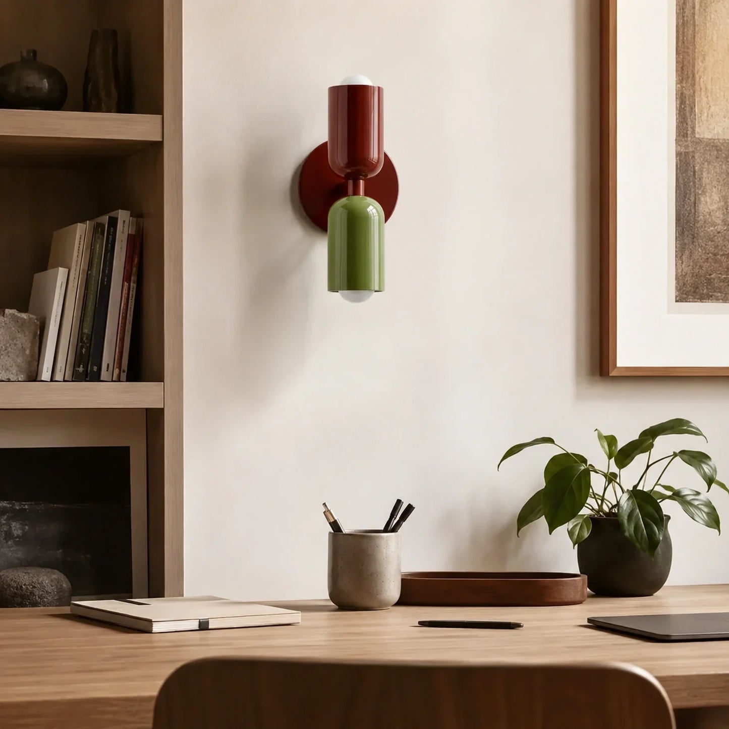 Contemporary dual-cylinder metal wall lamp in red wine and green mounted on a neutral wall above a wooden desk with a houseplant and stationery.