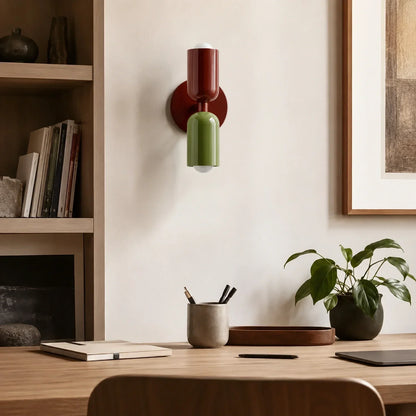 Contemporary dual-cylinder metal wall lamp in red wine and green mounted on a neutral wall above a wooden desk with a houseplant and stationery.
