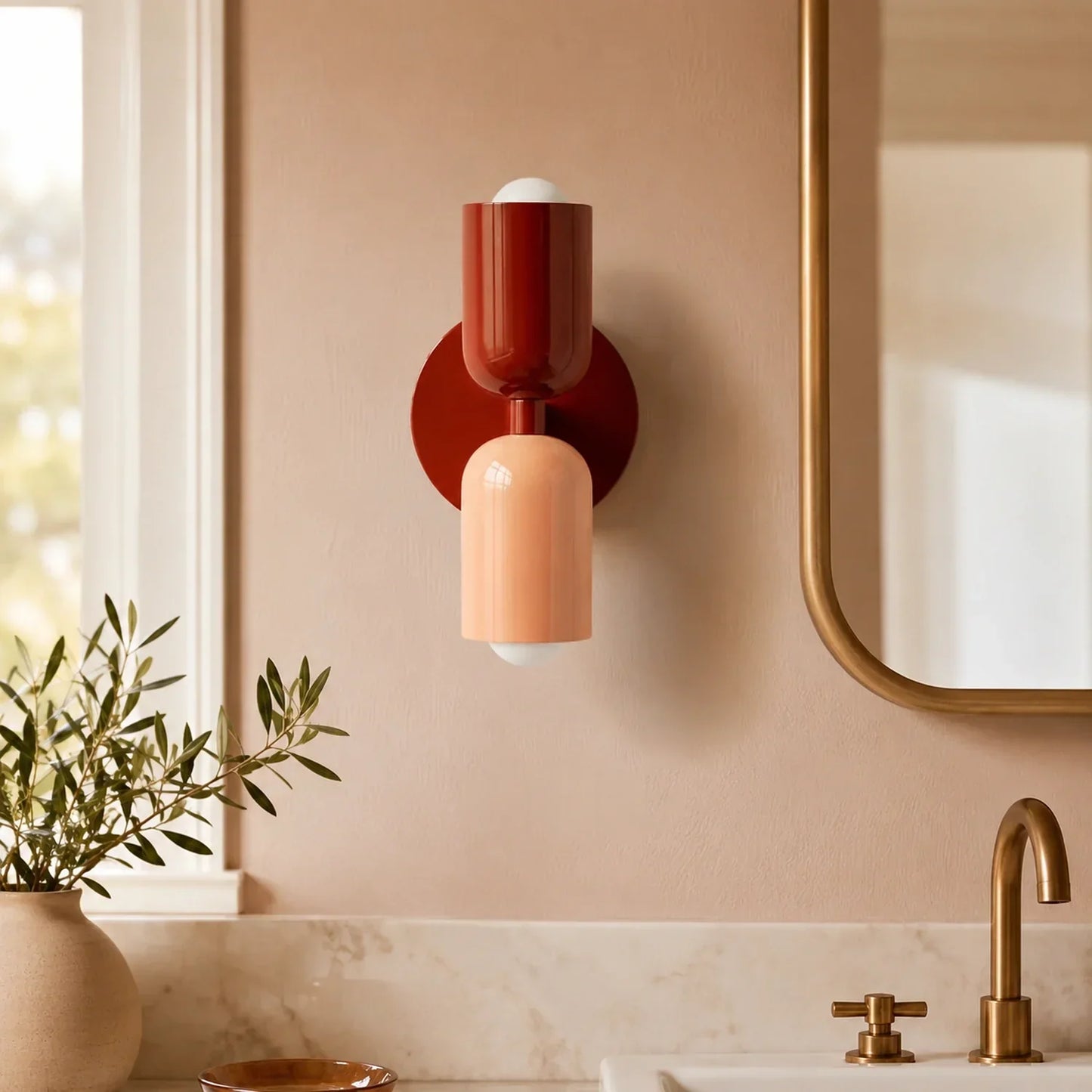 Contemporary dual-cylinder metal wall lamp in red wine and peach tones, mounted on a neutral bathroom wall above a marble vanity with brass fixtures.