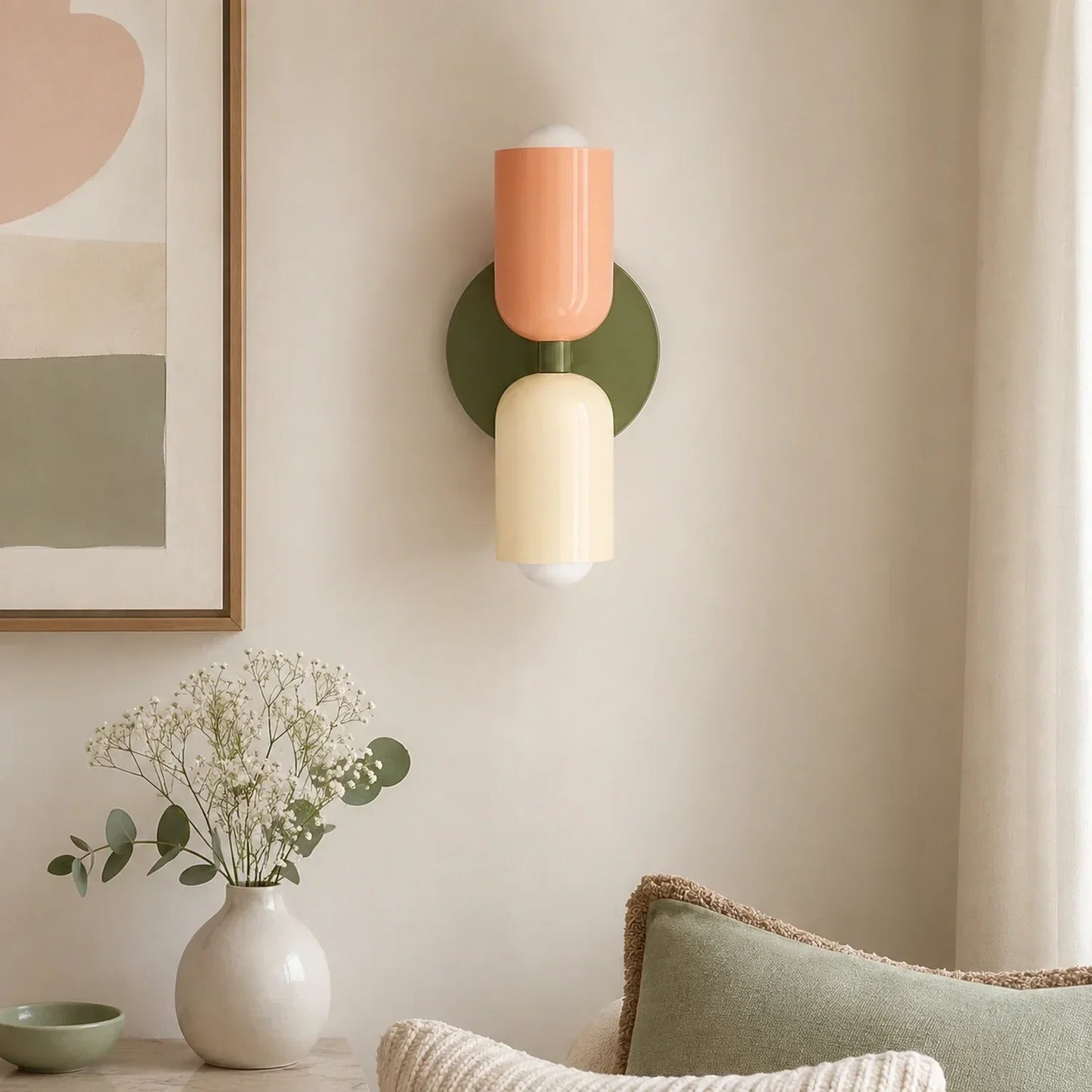 Contemporary dual-cylinder wall lamp in green, peach and cream mounted on a neutral wall above a vase of flowers in a modern living room interior.