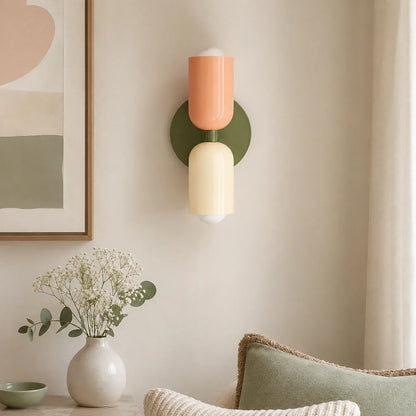 Contemporary dual-cylinder wall lamp in green, peach and cream mounted on a neutral wall above a vase of flowers in a modern living room interior.