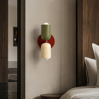 Contemporary dual-cylinder wall lamp featuring a red wine circular base with green and cream cylindrical shades mounted on a neutral bedroom wall.