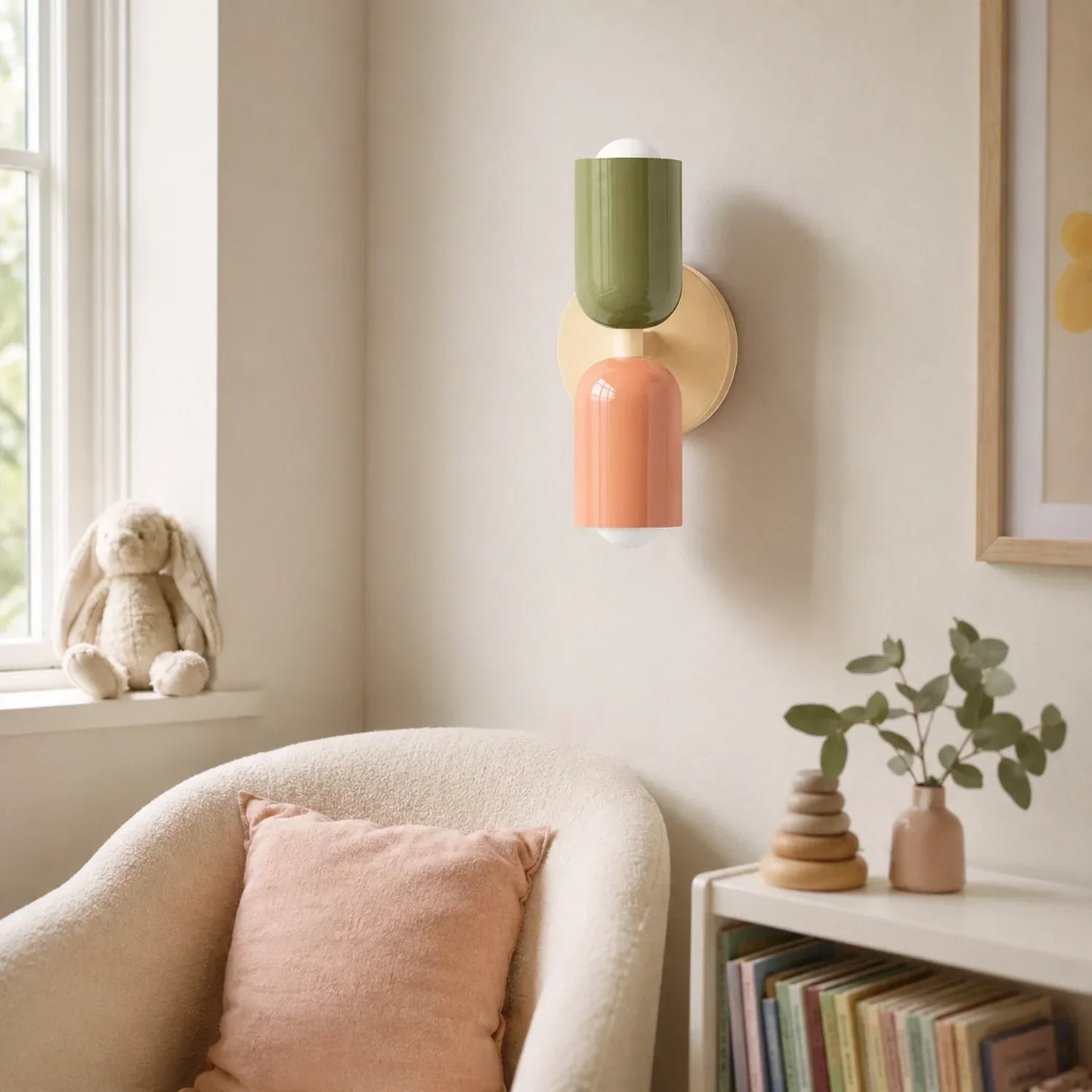 Contemporary dual-cylinder metal wall lamp with green and peach shades mounted on a beige wall in a cosy nursery with a soft armchair and bookshelf.