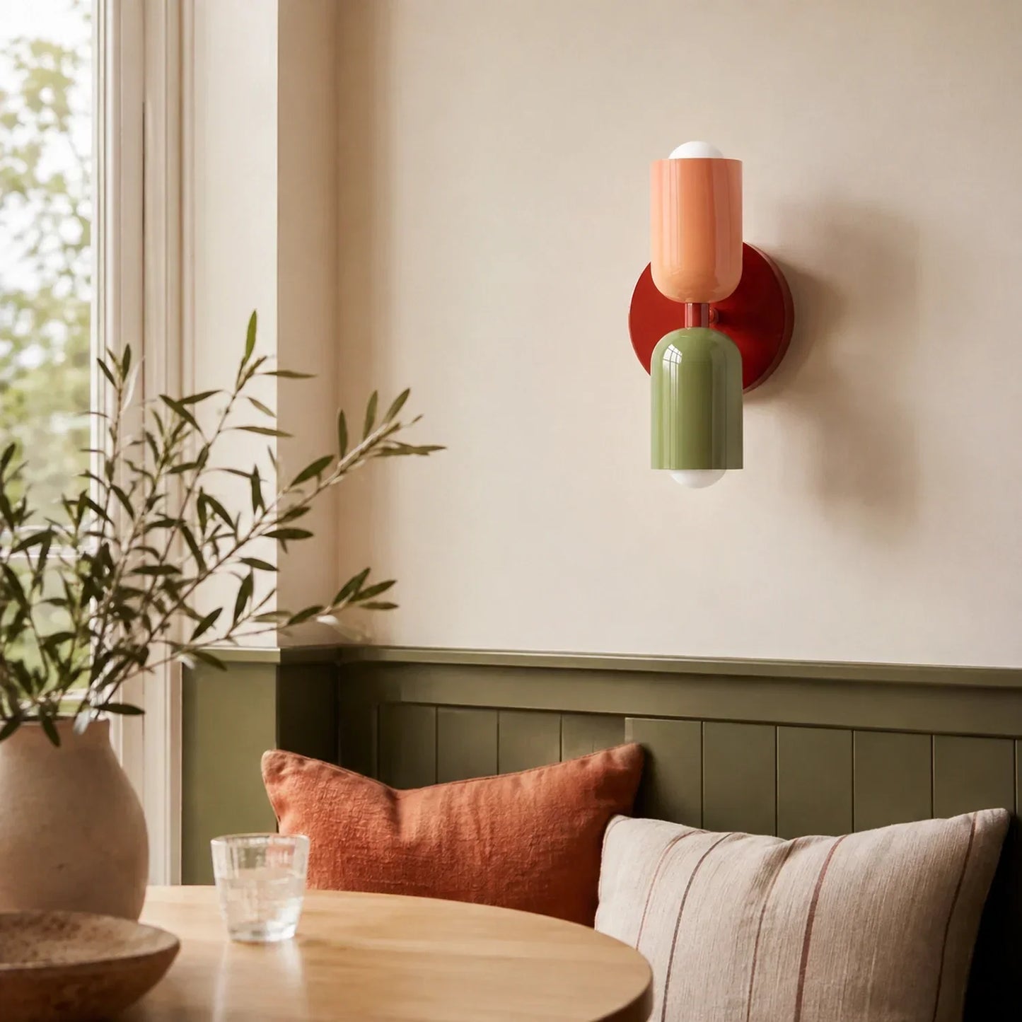 Contemporary dual-cylinder metal wall lamp with a red wine base, peach top shade and green bottom shade, mounted on a beige wall above a dining nook.