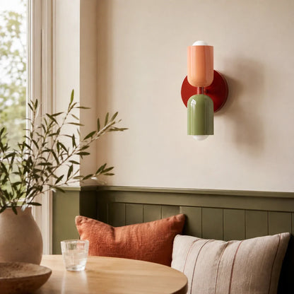 Contemporary dual-cylinder metal wall lamp with a red wine base, peach top shade and green bottom shade, mounted on a beige wall above a dining nook.