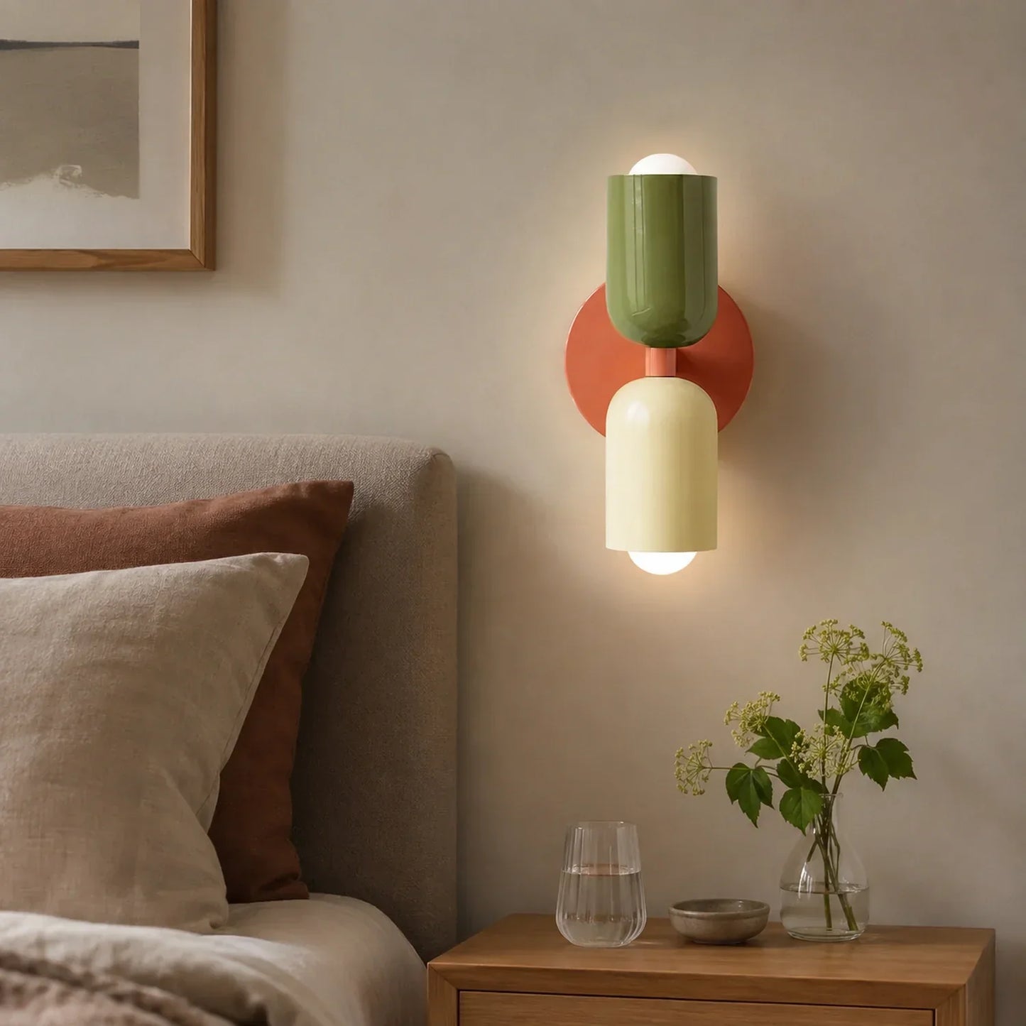 Contemporary dual-cylinder metal wall lamp with a pink circular base, green upper shade and pale yellow lower shade, mounted above a bedroom bedside table.