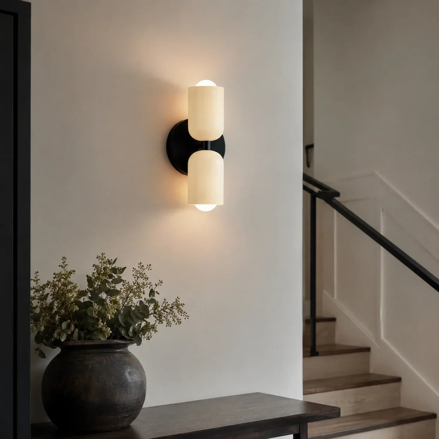 Contemporary black dual-cylinder wall lamp with glowing shades mounted on a neutral wall above a console table with a decorative plant and staircase.