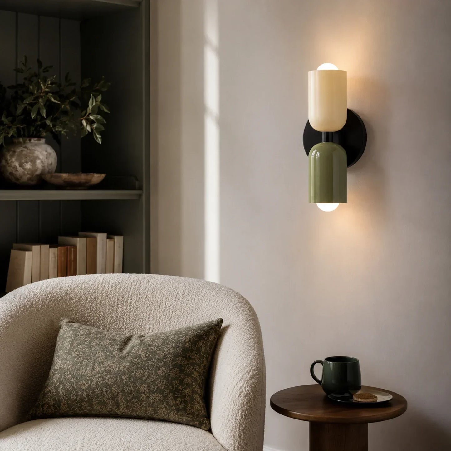 Contemporary dual-cylinder wall lamp with cream and olive green shades mounted on a neutral wall above a textured armchair in a modern living room.