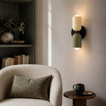 Contemporary dual-cylinder wall lamp with cream and olive green shades mounted on a neutral wall above a textured armchair in a modern living room.