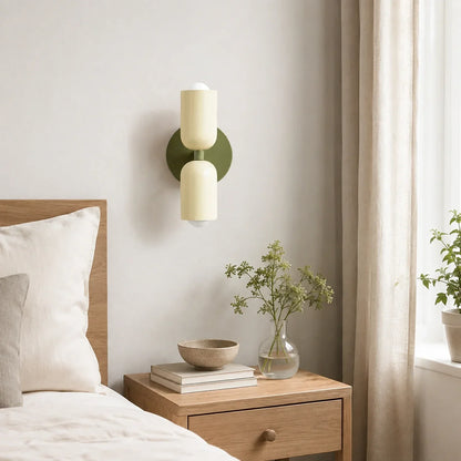 Contemporary dual-cylinder metal wall lamp with a green base and cream shades, mounted above a wooden bedside table in a modern, neutral bedroom.