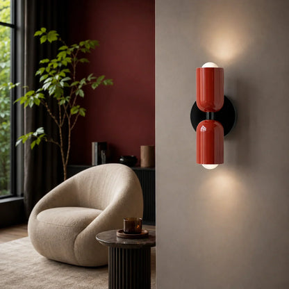 Glossy red dual-cylinder wall lamp mounted on a neutral wall, casting a warm up and down light in a contemporary living room with a cream armchair.