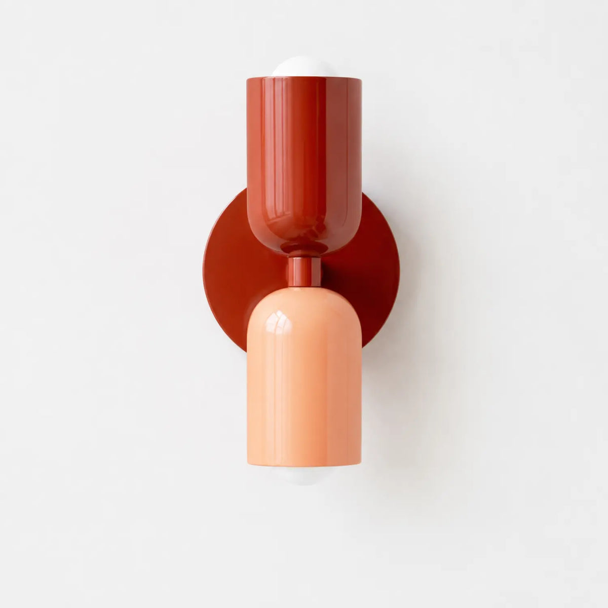 Contemporary dual-cylinder wall lamp featuring a deep red upper shade and a soft peach lower shade mounted on a circular red base against a white wall.