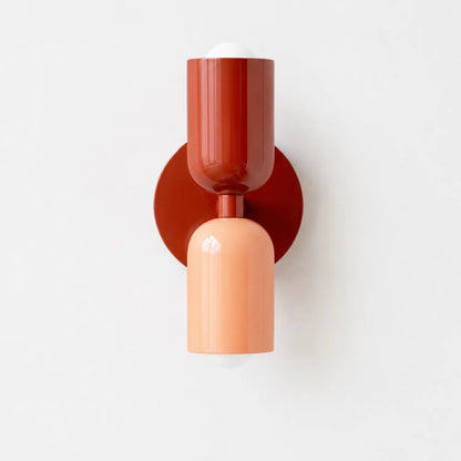 Contemporary dual-cylinder wall lamp featuring a deep red upper shade and a soft peach lower shade mounted on a circular red base against a white wall.