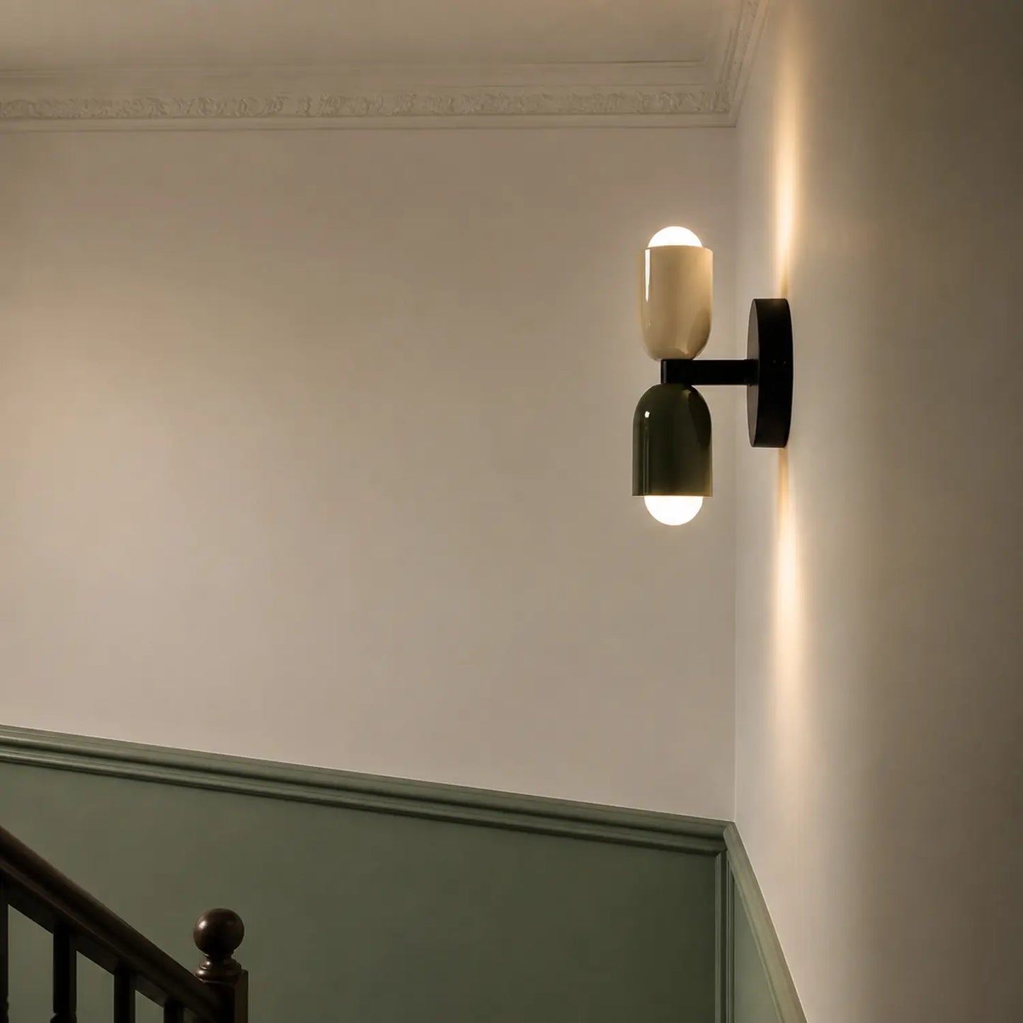Contemporary dual-cylinder wall lamp with cream and green shades mounted on a white wall above a green panelled staircase, casting a warm up-down glow.