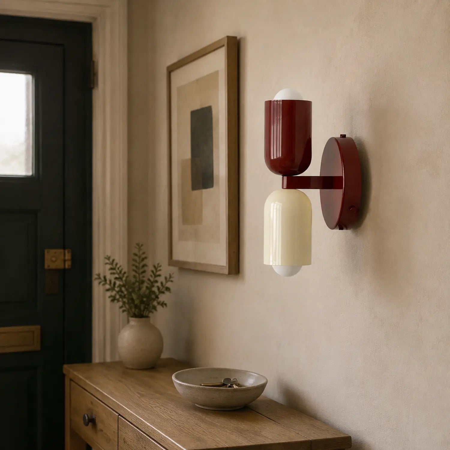 Contemporary dual-cylinder metal wall lamp with red and cream shades mounted on a neutral hallway wall above a wooden console table with decor.