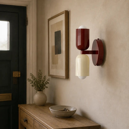 Contemporary dual-cylinder metal wall lamp with red and cream shades mounted on a neutral hallway wall above a wooden console table with decor.