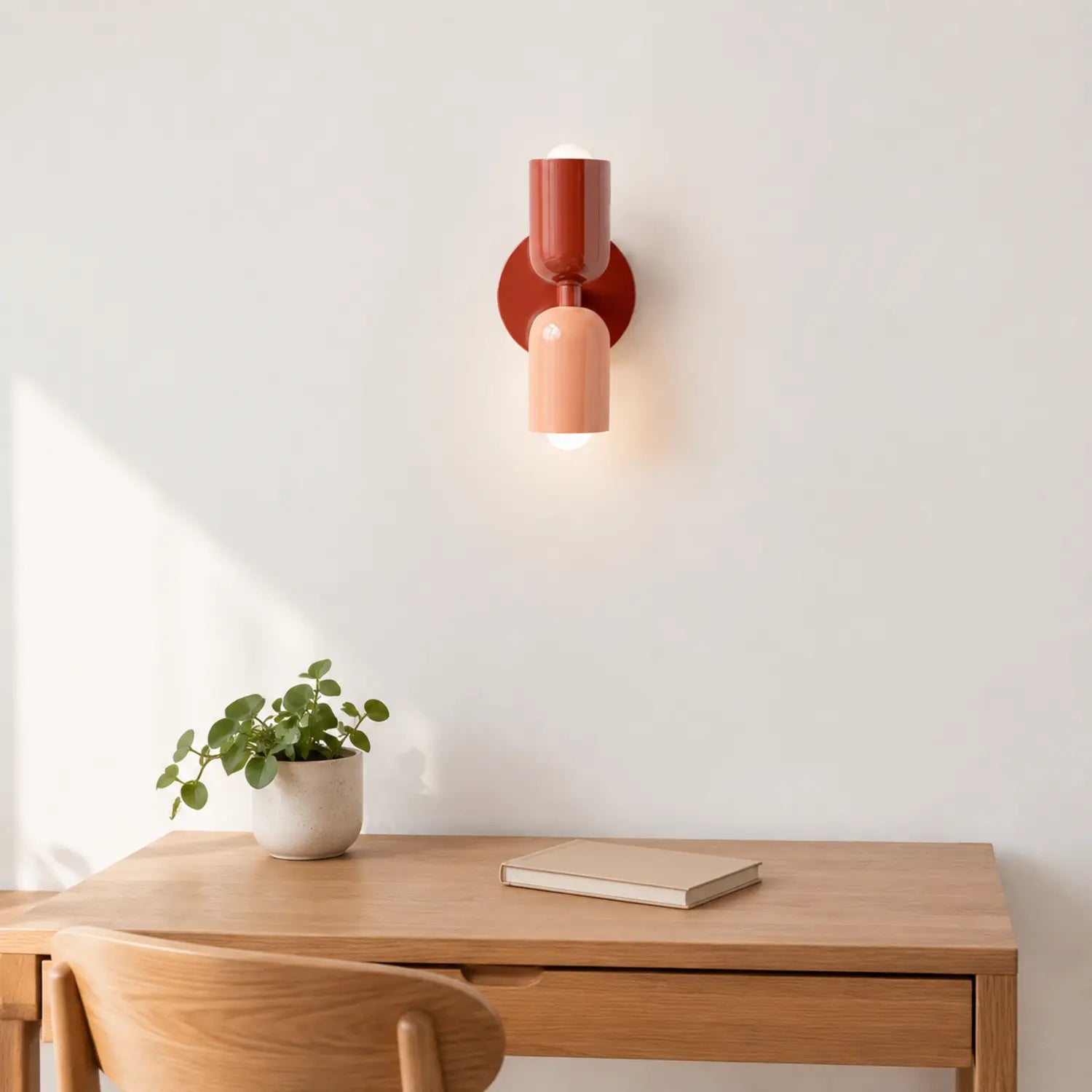 Contemporary dual-cylinder metal wall lamp in rust and peach mounted above a wooden desk with a small potted plant and a closed book in a home office.