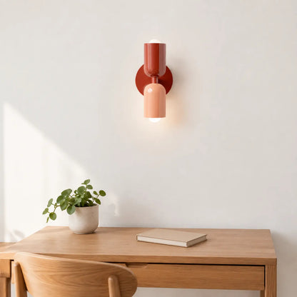 Contemporary dual-cylinder metal wall lamp in rust and peach mounted above a wooden desk with a small potted plant and a closed book in a home office.