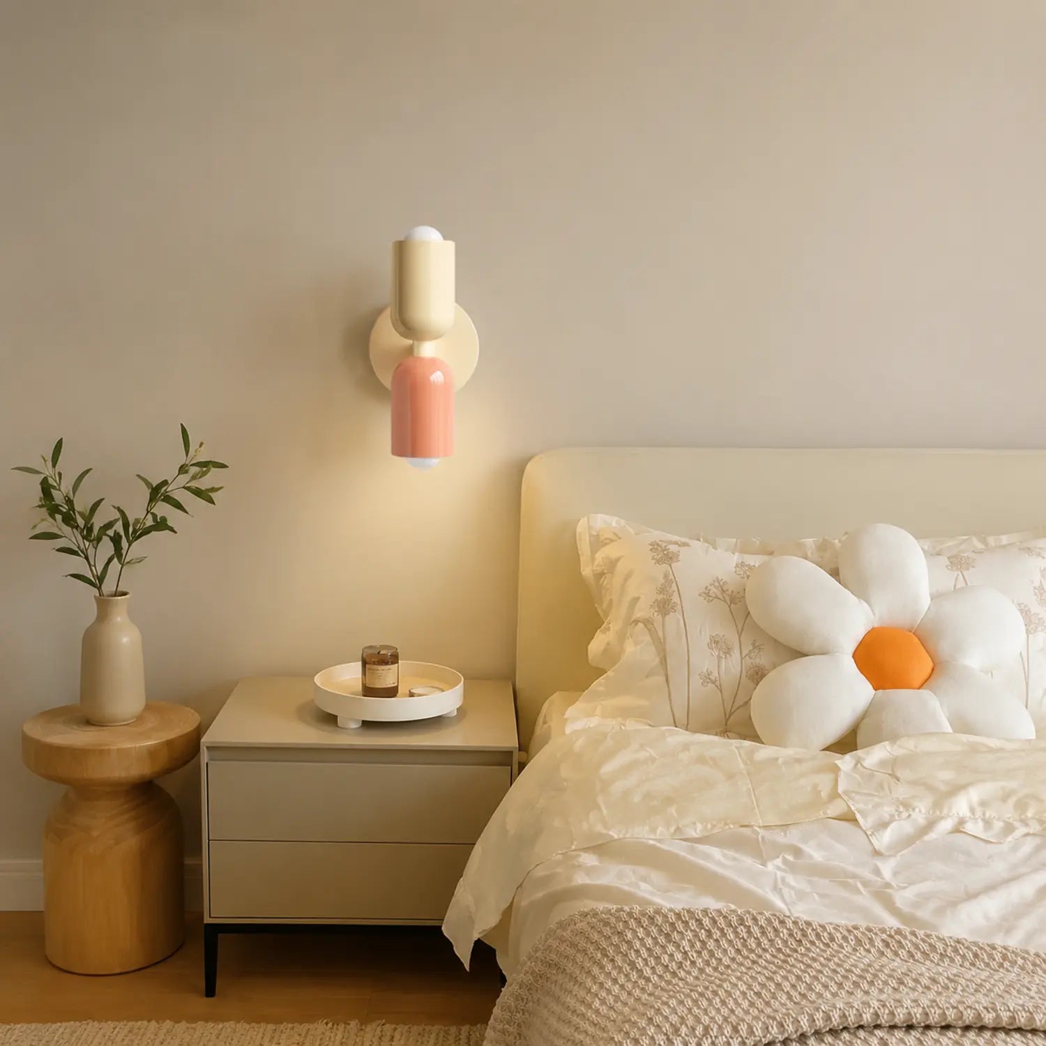 Contemporary dual-cylinder metal wall lamp in cream and pink mounted above a bedside table in a modern bedroom with neutral decor and floral accents.