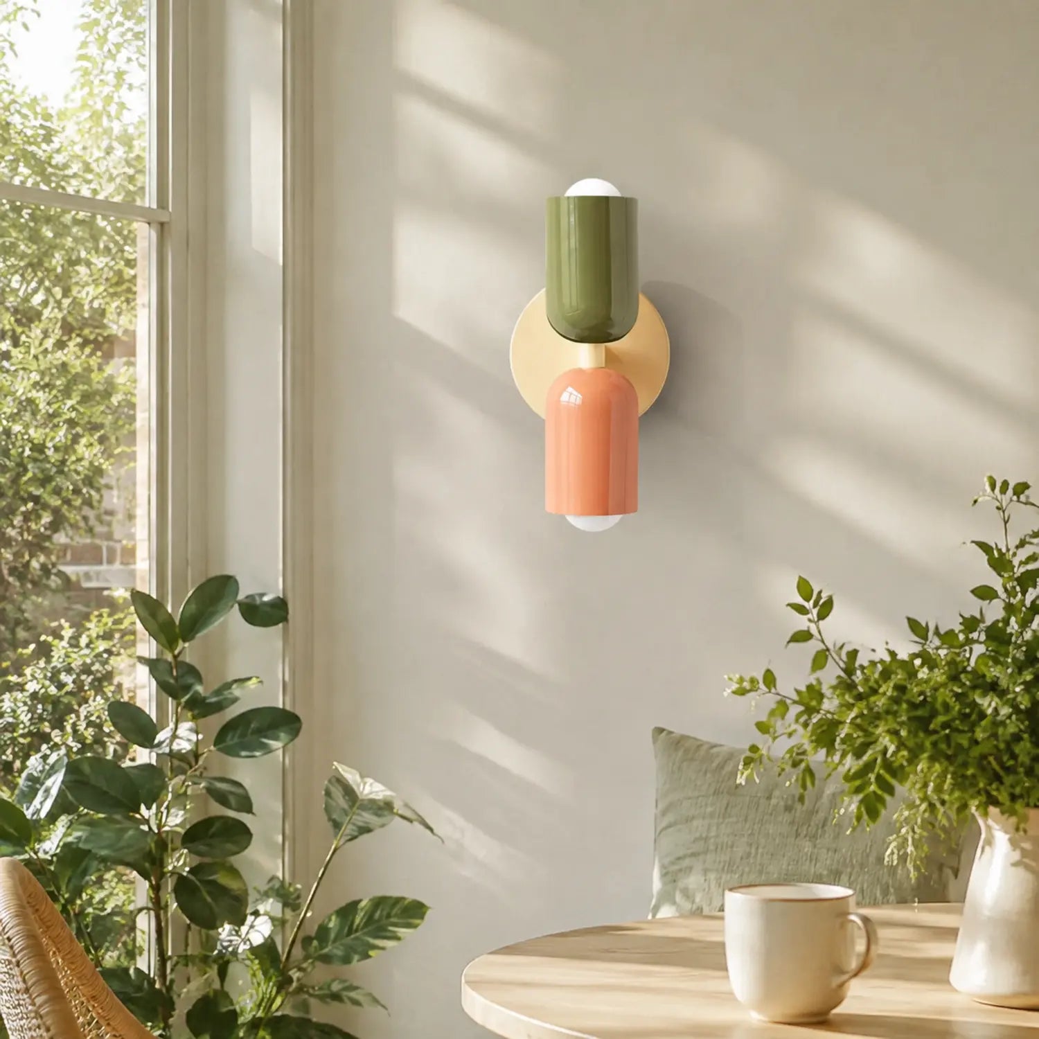 Contemporary dual-cylinder metal wall lamp with green and orange shades mounted on a neutral wall in a sunlit room with indoor plants and a table.