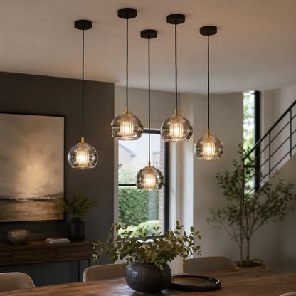 Five contemporary ribbed glass globe pendant lights with brass accents suspended at varying heights above a wooden dining table in a modern home interior.