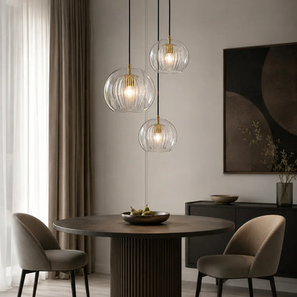 Three contemporary ribbed glass globe pendant lights with brass fittings hanging above a round dining table in a modern, neutral-toned dining room.