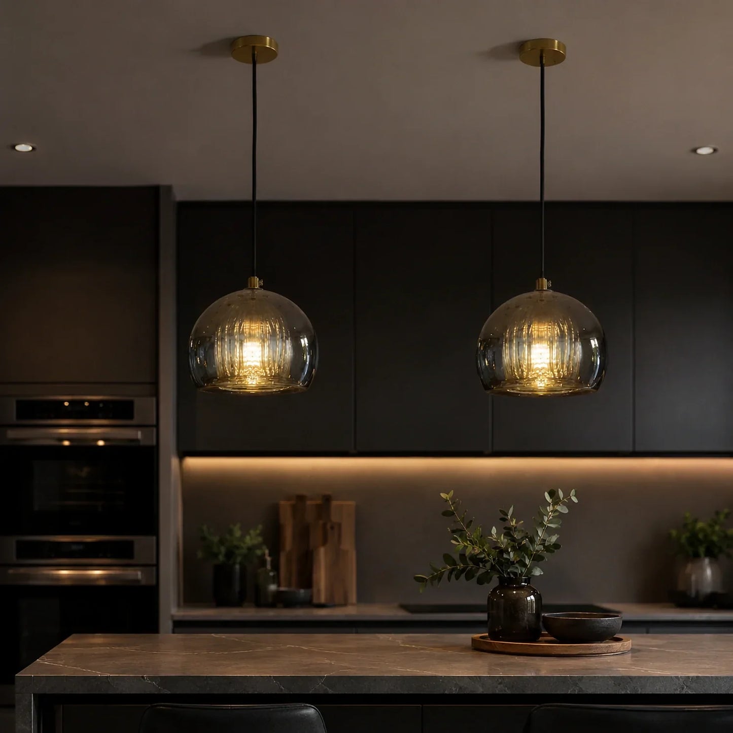 Two contemporary ribbed glass globe pendant lights with warm glowing bulbs hanging above a dark modern kitchen island with a decorative plant.