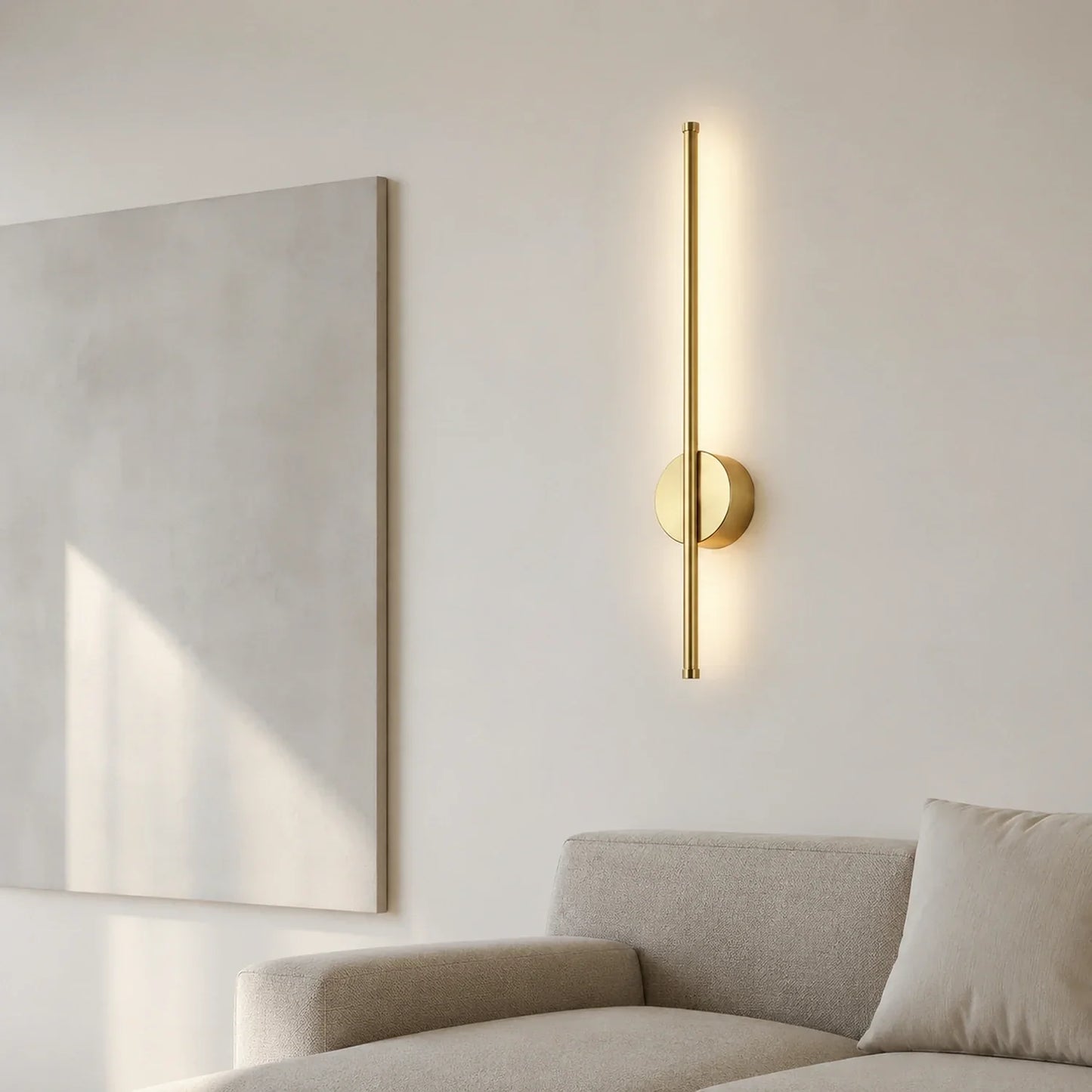 A modern, vertical gold LED wall light mounted on a neutral wall above a sofa, emitting a warm, soft glow in a contemporary living room setting.