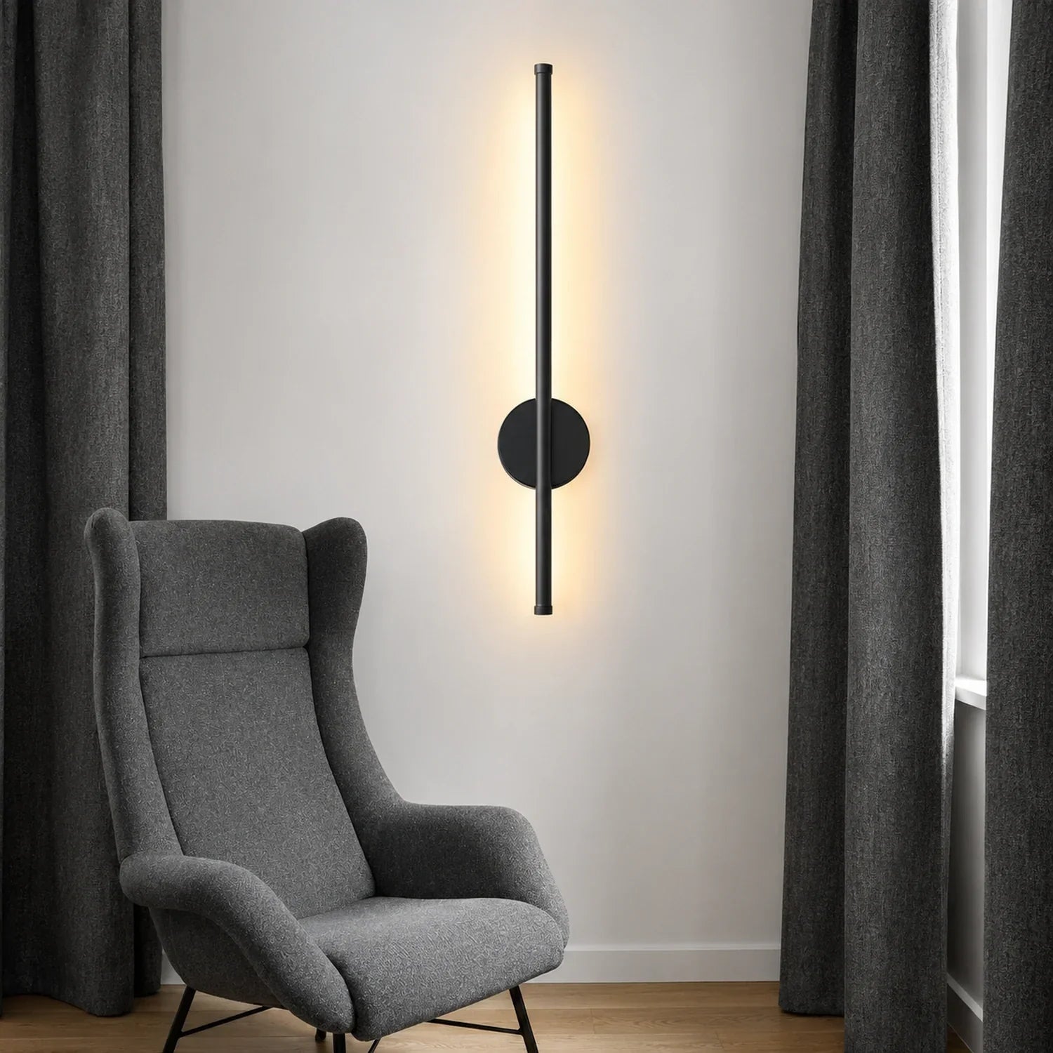 Modern black vertical LED wall light mounted on a white wall above a grey armchair, casting a warm glow in a contemporary living room setting.