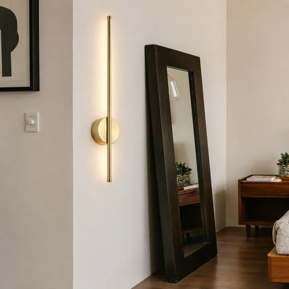 Modern gold-toned linear LED wall light mounted on a white bedroom wall, featuring a slim vertical design that provides soft up and down illumination.