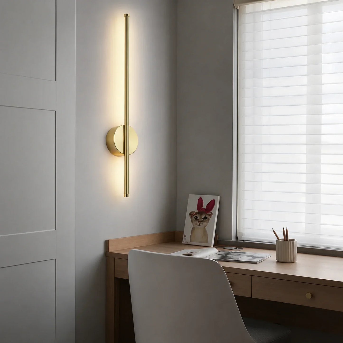 A modern gold vertical LED wall light mounted above a wooden desk in a bedroom, casting a warm glow on the grey wall next to a window with blinds.
