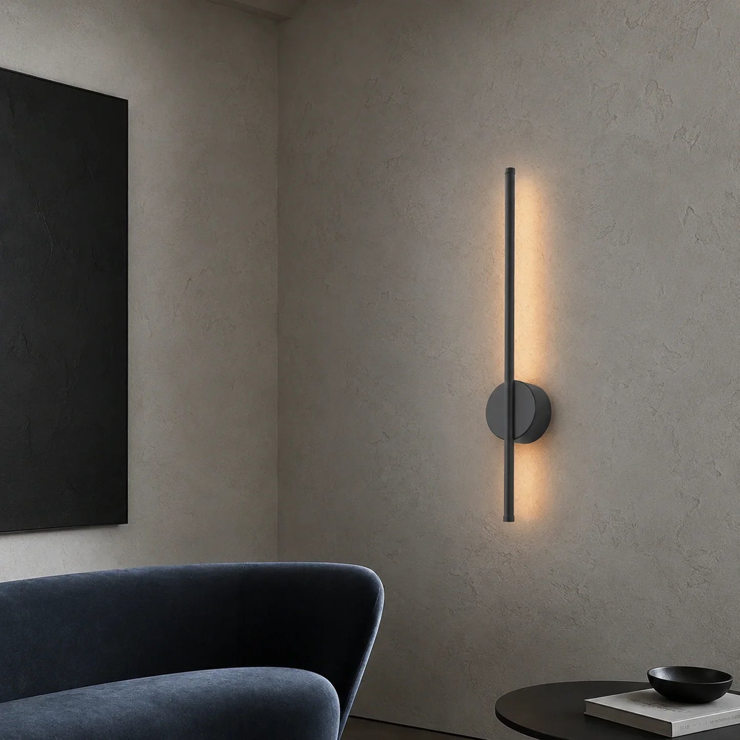 A sleek, modern black vertical LED wall light mounted on a textured grey wall, casting a warm, soft glow in a contemporary living room setting.