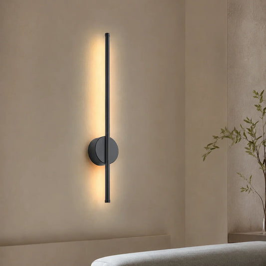 A modern, slimline black LED wall light mounted vertically, emitting a warm, soft glow against a neutral bedroom wall for elegant ambient lighting.