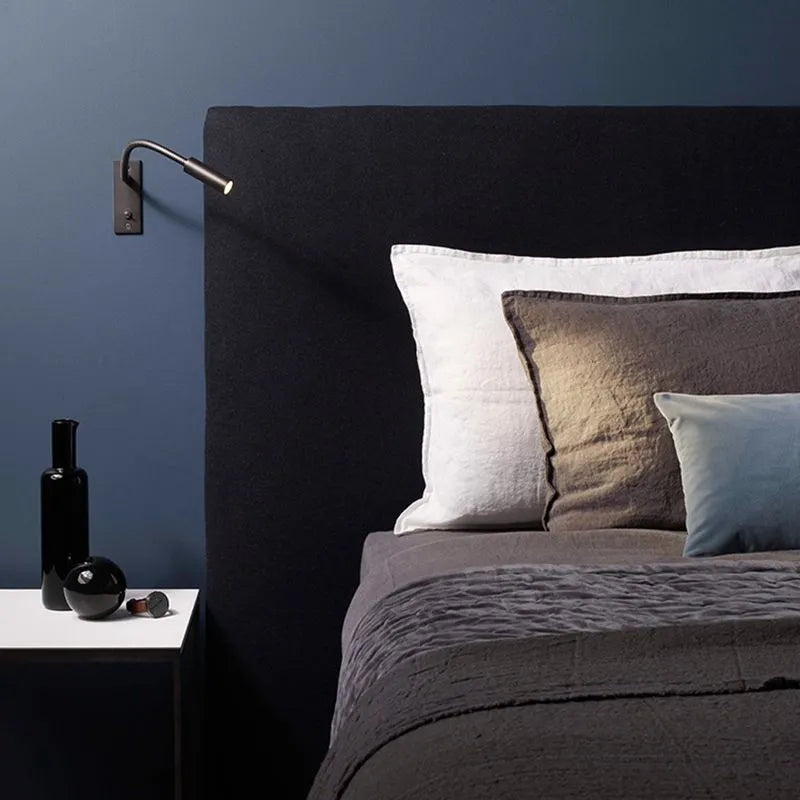 A flexible LED reading wall lamp mounted above a dark upholstered headboard in a modern bedroom, providing focused light for bedside reading.