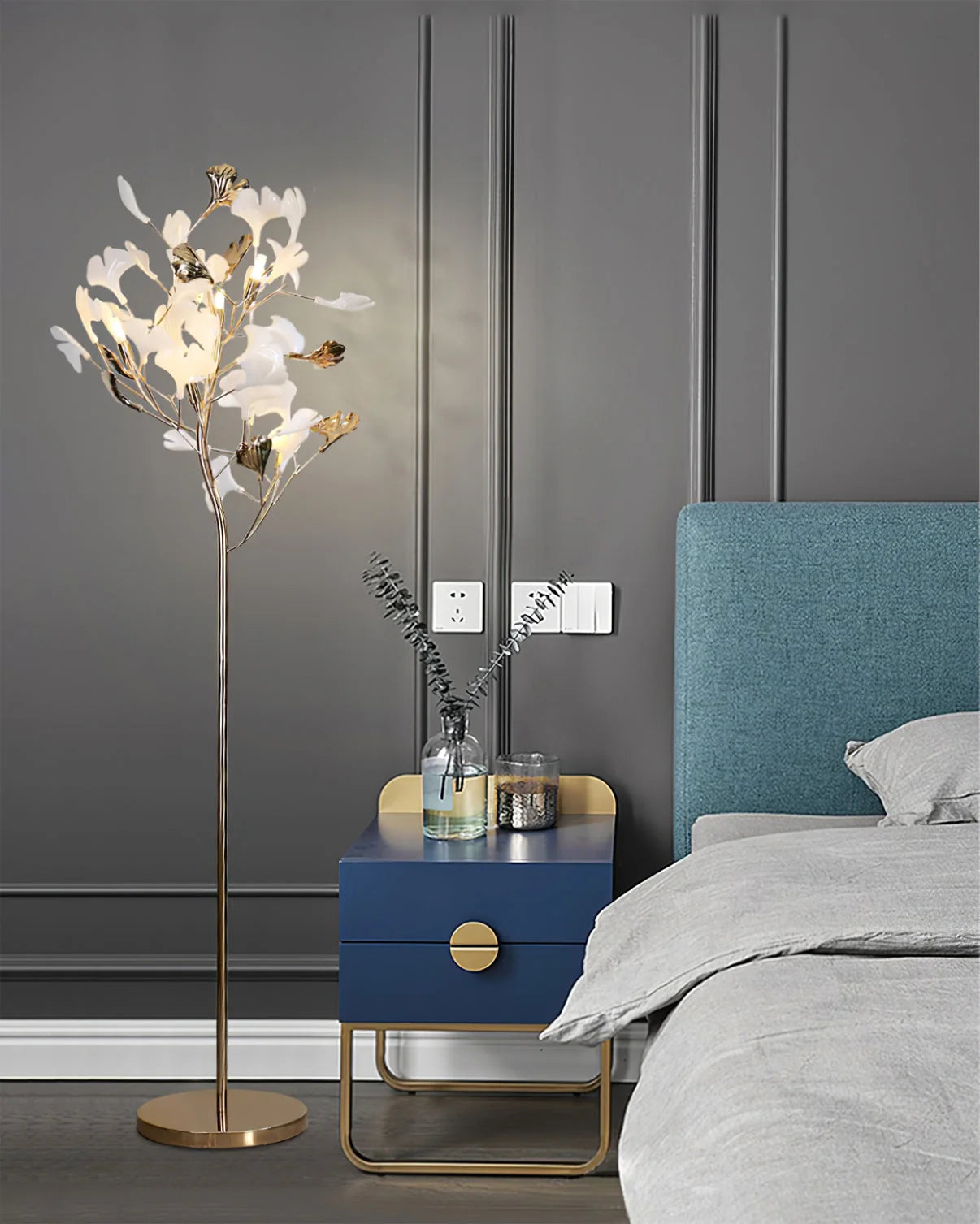 An illuminated gold Ginkgo Leaf LED floor lamp standing next to a blue bedside table in a modern bedroom with a grey panelled wall.
