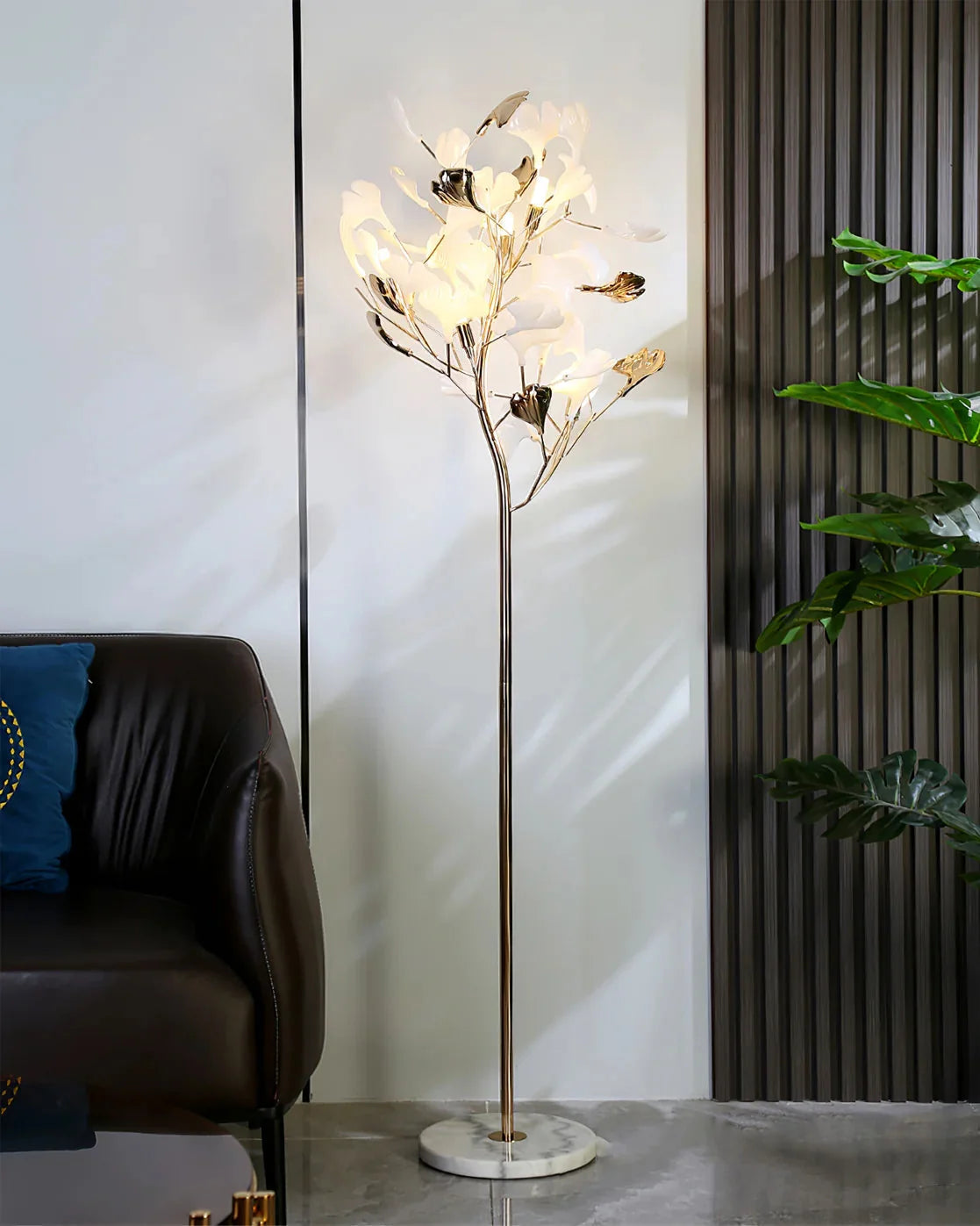 Elegant Ginkgo Leaf LED floor lamp with a gold-coloured stem and marble base, illuminated in a modern living room next to a leather armchair.