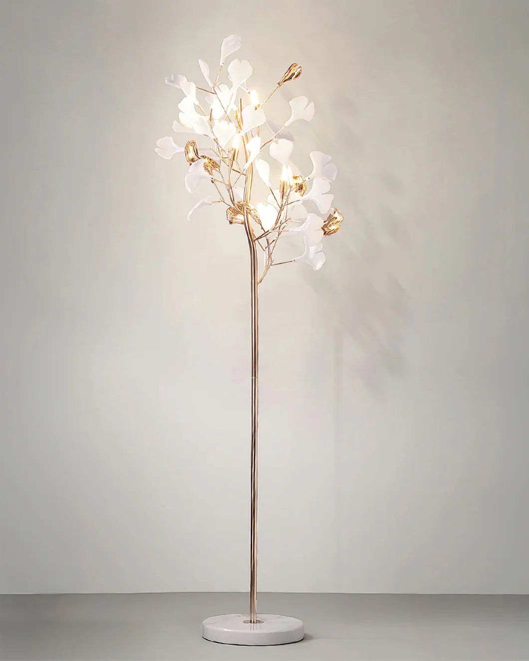 A gold Ginkgo Leaf LED floor lamp with five glowing lights and white leaf-shaped shades, standing on a round white marble base against a plain wall.