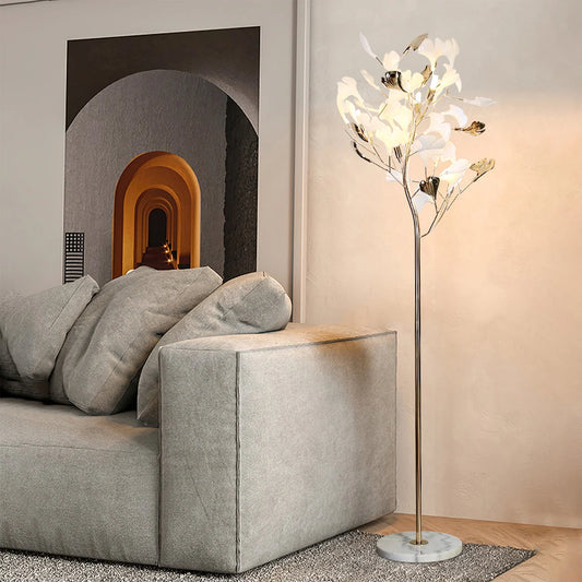 A modern ginkgo leaf LED floor lamp with a marble base and warm glowing lights, standing next to a grey sofa in a contemporary living room.
