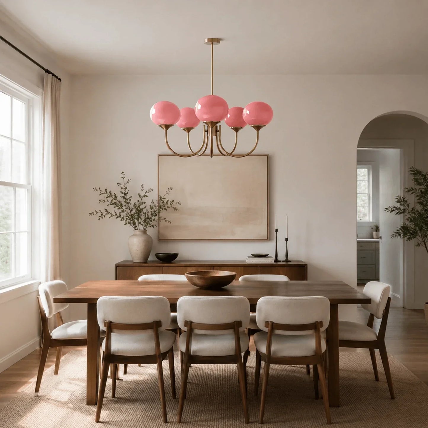 A modern brass pendant light with five pink glass ball globes suspended above a wooden dining table in a stylish, neutral-toned dining room.