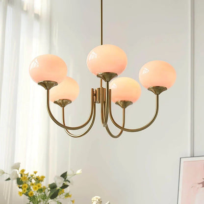 Modern five-light pendant chandelier with curved brass arms and glowing peach-pink glass ball shades, hanging in a bright, minimalist room.