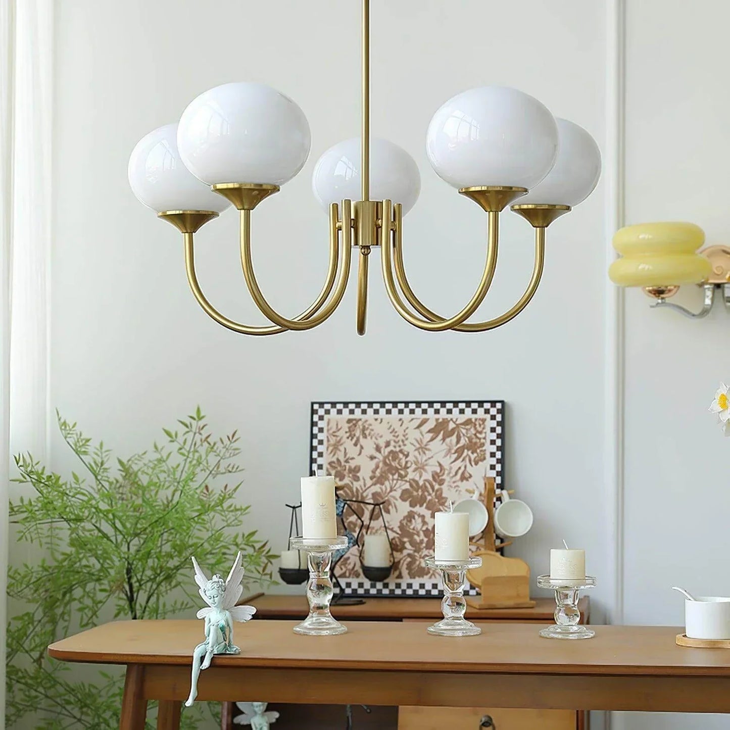 A modern five-light brass pendant ceiling fixture with white glass ball globes, hanging in a dining room above a wooden table with candles.