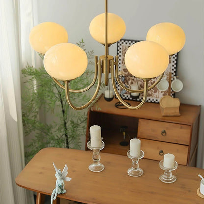 Modern five-light pendant ceiling fixture featuring glowing frosted glass ball globes on a curved brass metal frame, hanging above a wooden table.
