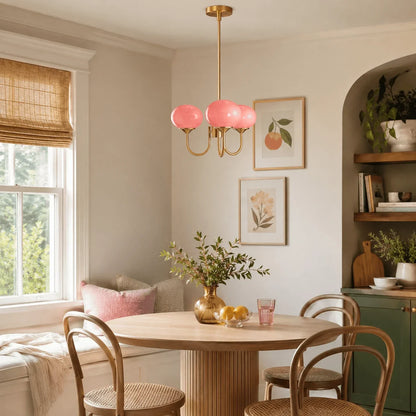 A modern brass pendant light with three pink glass ball globes hanging above a wooden dining table in a cosy, bright dining room.