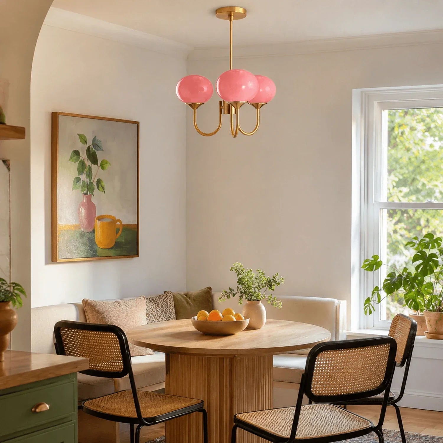 A modern three-light pendant ceiling light with pink glass globes and a brass metal finish, hanging above a wooden dining table in a bright room.
