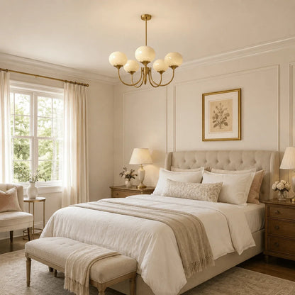 A modern glass ball pendant light with a brass finish hanging in a neutral-toned bedroom with a tufted headboard and elegant interior decor.