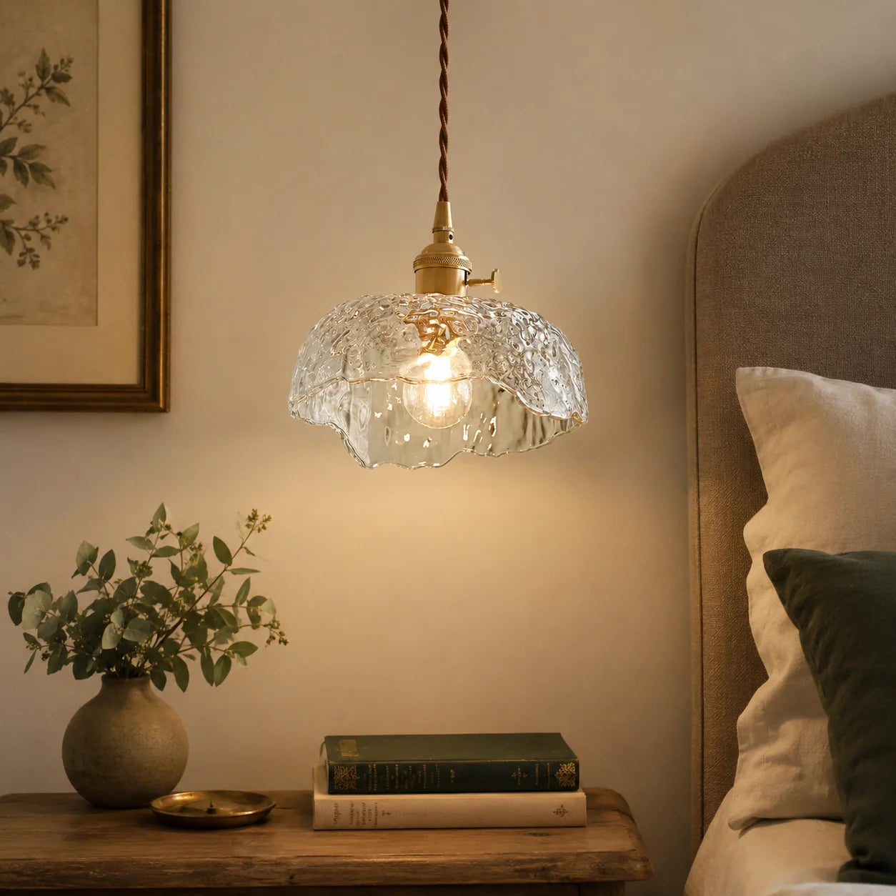 Modern glass pendant light with a textured shade and brass fitting, suspended by a cord above a wooden bedside table in a cosy bedroom setting.